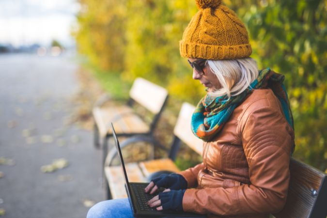 Mulher sentada em um banco de um parque com roupa de frio e trabalhando na modalidade anywhere office